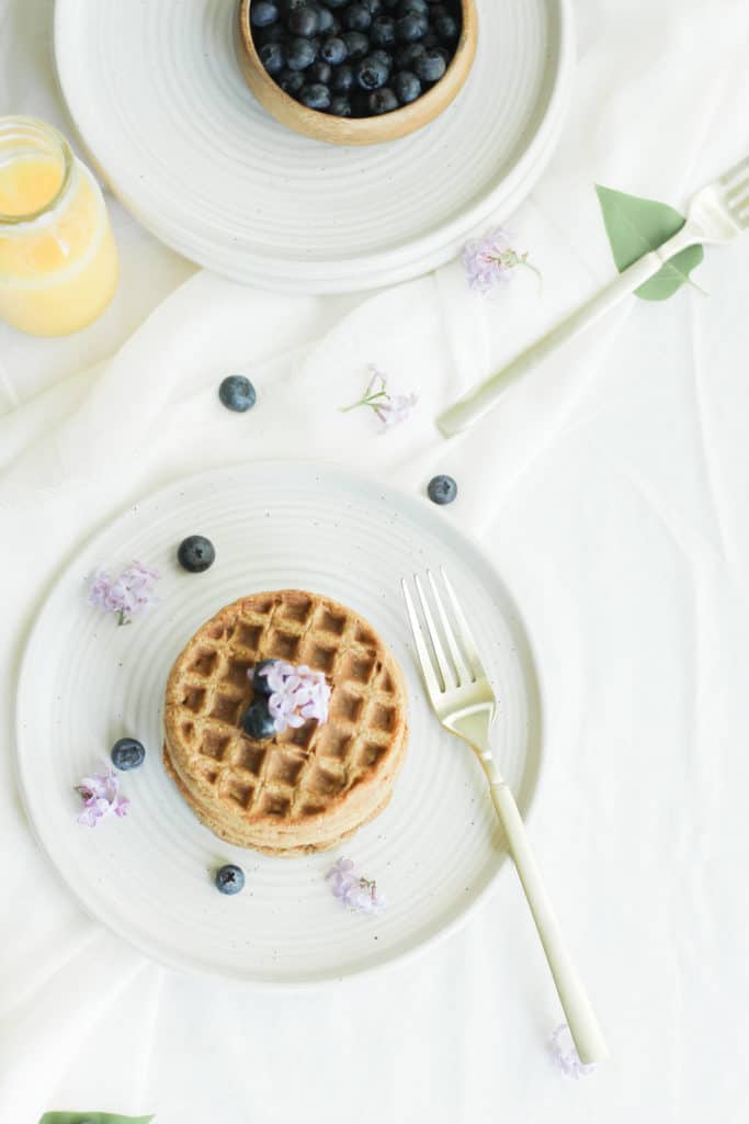 Overhead picture of waffles on a white plate with blueberries and a gold fork. Flowers and orange juice decorate the scene