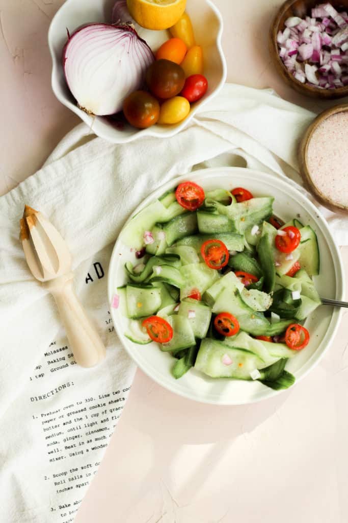 Cucumber salad in a white bowl on a pink backdrop. vegetables and a lemon juicer surround it