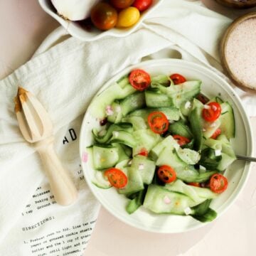 cucumber salad in a white bowl with red onion, sliced tomatoes, and a lemon juicer on the side.