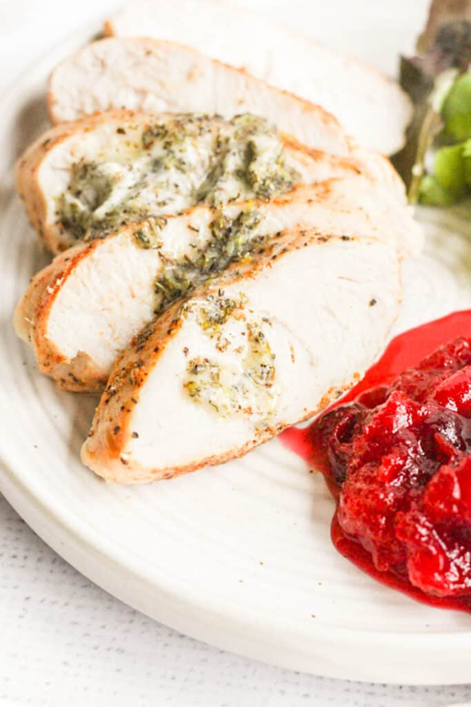 healthy turkey breast cooked with herb butter melting over top of it on white plate with cranberry sauce.