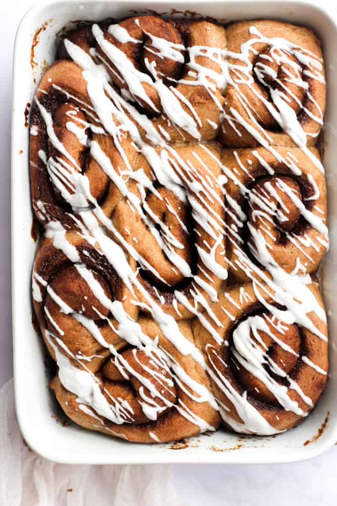 healthier whole wheat cinnamon rolls on a white baking dish drizzled with a vanilla glaze