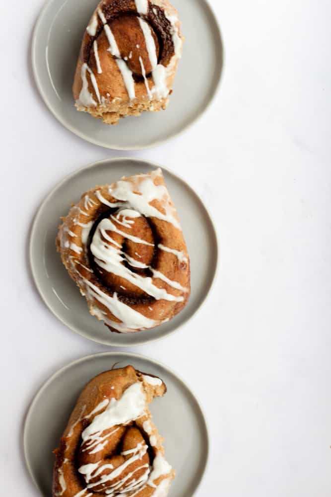 three whole wheat cinnamon rolls on separate gray plates in a vertical line on a white backdrop.