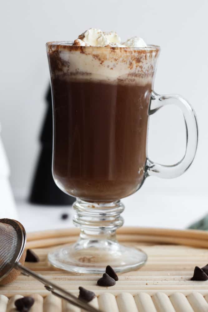 clear glass mug full of healthy sugar free hot cocoa on a wood board. it is topped with whipped cream and cocoa powder