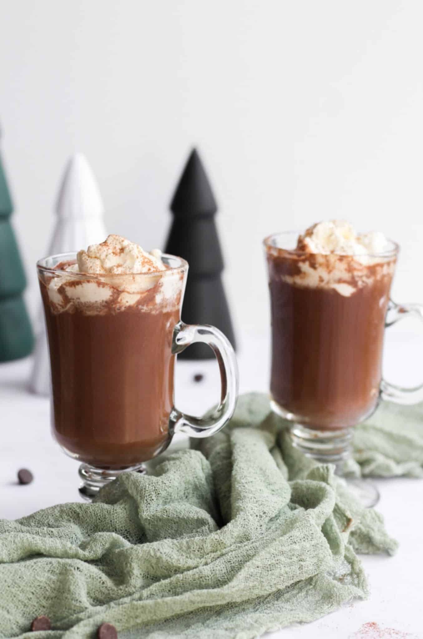 two glass mugs of sugar free hot chocolate topped with whipped cream and a green napkin between them