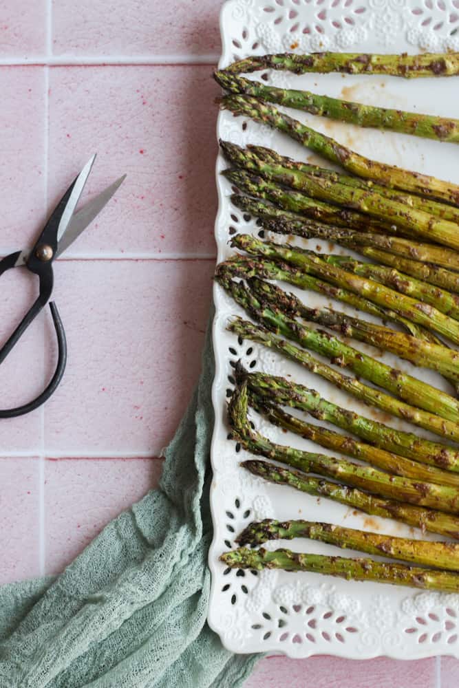 air fryer balsamic asparagus on a white platter on a pink backdrop with a pair of Chinese flower scissors to the left of the platter