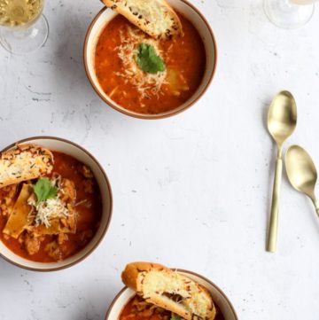 gluten free lasagna soup in three white bowls topped with toasted bread and extra cheese on a white backdrop