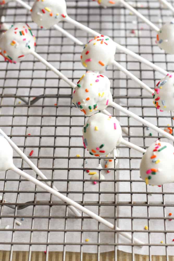 gluten free cake pops dipped in white chocolate and sprinkles cooling on a cooling rack over parchment paper