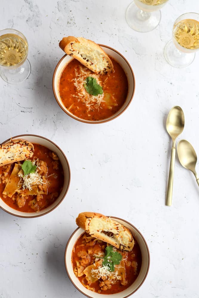 gluten free lasagna soup in three bowls in a white backdrop topped with toasted bread and cheese