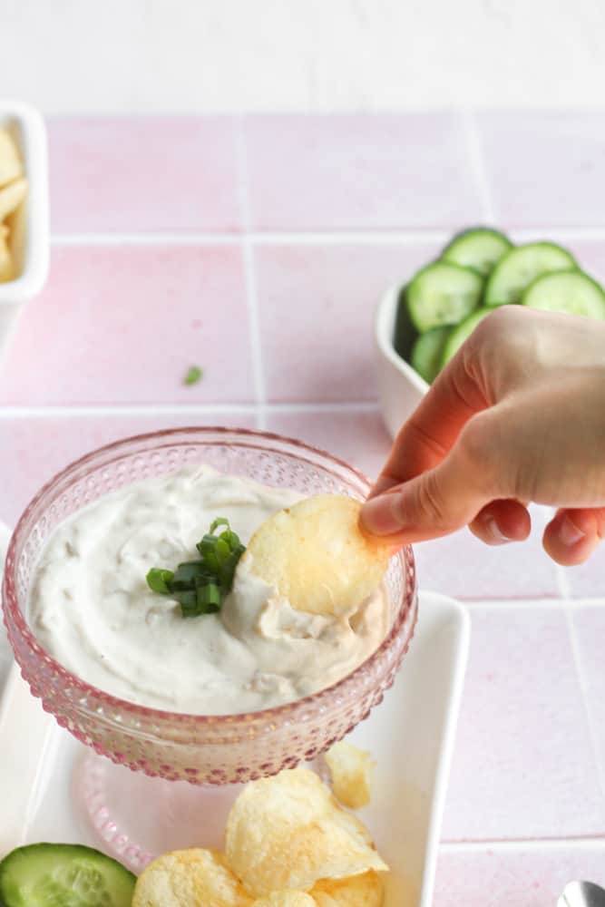 hand dipping a potato chip into Greek yogurt dip in a pink glass bowl photographed on a pick backdrop