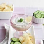 healthy French onion dip in a pink glass bowl on a white platter with sliced cucumbers and potato chips