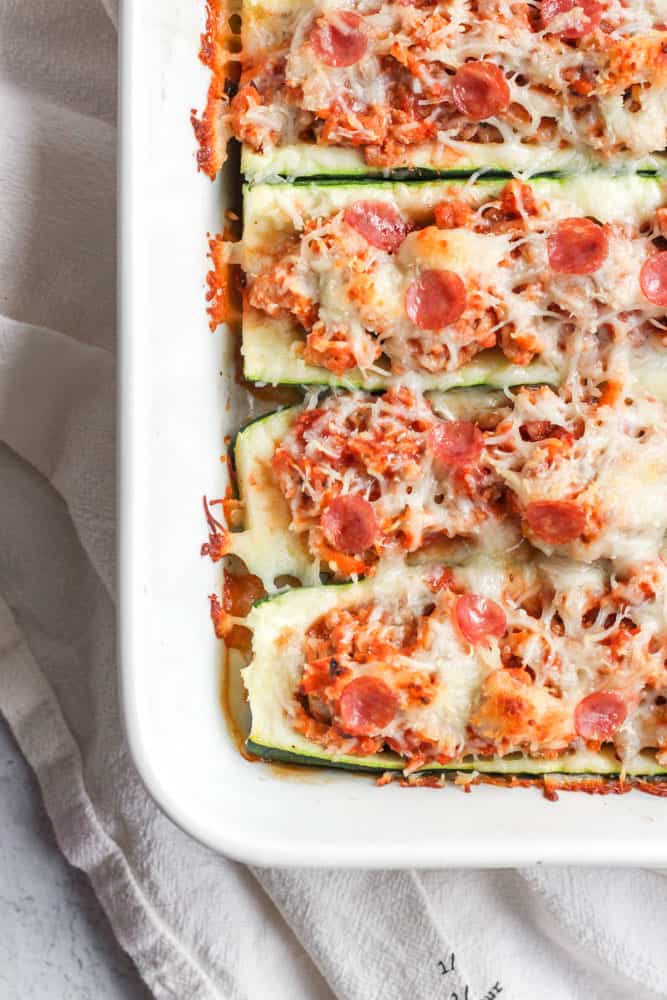 healthy zucchini pizza boats in a white baking dish. the boats are covered in melted mozzarella cheese and mini pepperoni
