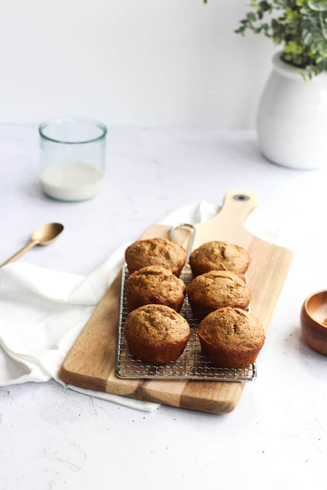 6 healthy, easy banana bread muffins on a silver cooling rack resting on a wooden cutting board on a white backdrop. There's a white napkin underneath the wood board as well as a gold spoon to the left of it