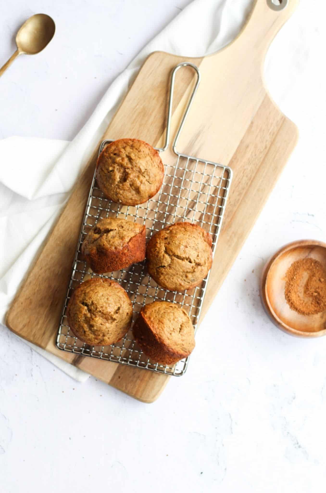 healthy 5 ingredient banana bread muffins on a cooling rack on top of a wooden cutting board on a white backdrop. A wooden bowl of coconut sugar is on the right hand side, and 2 of the muffins are turned on their sides