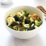 air fryer broccoli and cauliflower in a white bowl on a white backdrop with a dish of spices in the top left corner