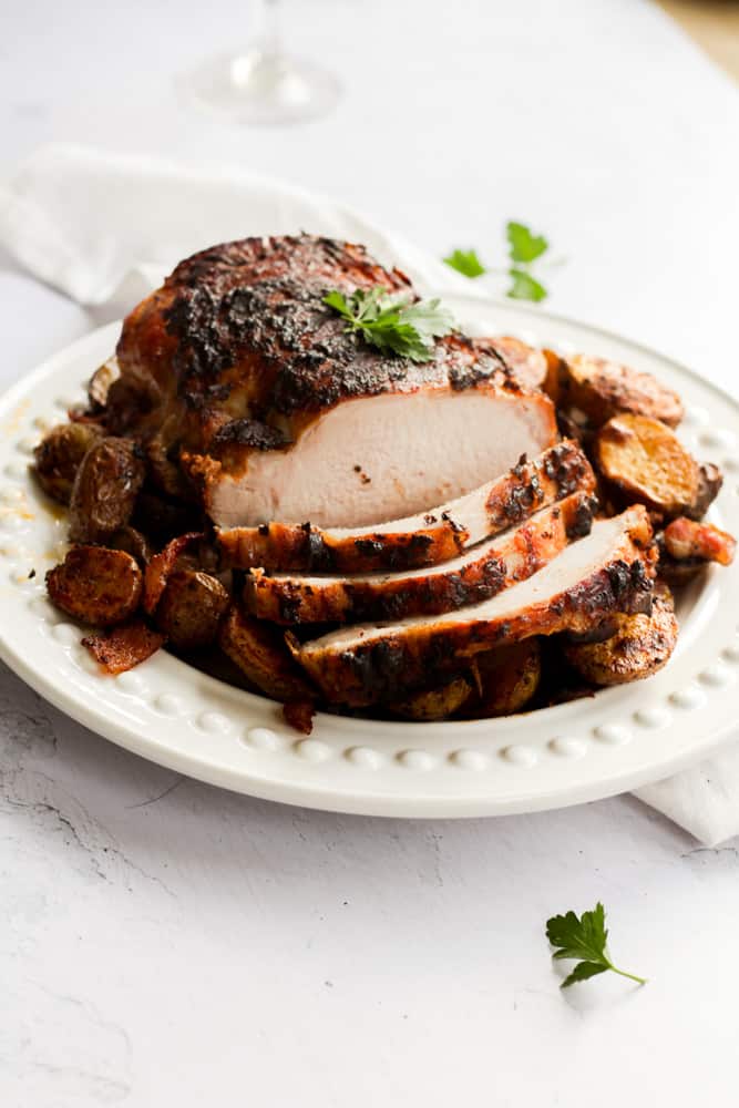 pork roast sliced and served over a bed of potatoes on a white platter. It is resting on a white backdrop with fresh parsley sprinkled around