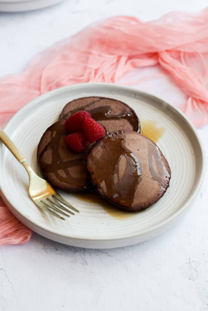 three vegan chocolate pancakes topped with syrup and raspberries on a white plate with a gold fork and pink napkin