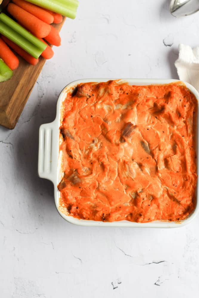 healthy dairy free chicken dip in a white baking dish on a white backdrop with cut up carrots and celery in the top left corner