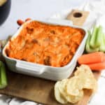 dairy free buffalo chicken dip baked in a white baking dish on a wood swuare board lined with carrots, celery, and potato chips on a whit backdrop