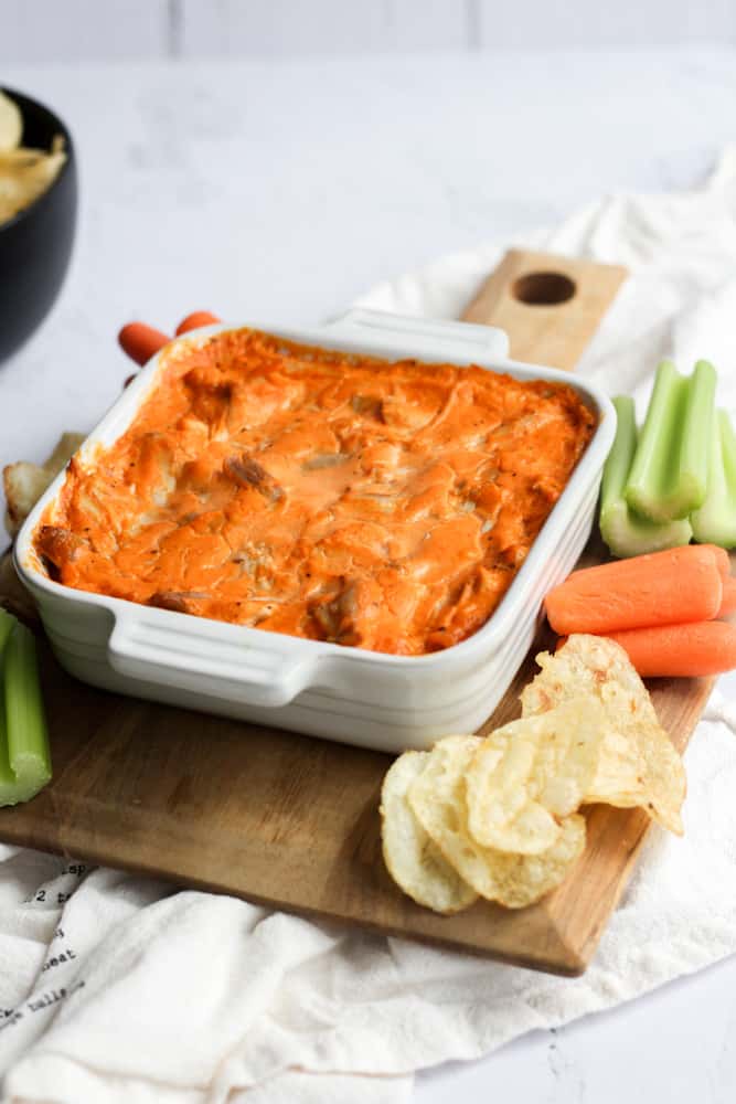 dairy free buffalo chicken dip baked in a white baking dish on a wood square board lined with carrots, celery, and potato chips on a whit backdrop