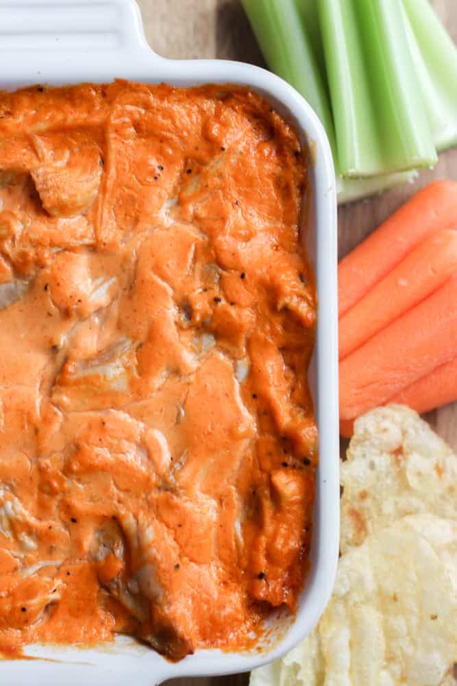 close up of healthy buffalo chicken dip in a white dish with potato chips and cut carrots and celery on the right side of the frame