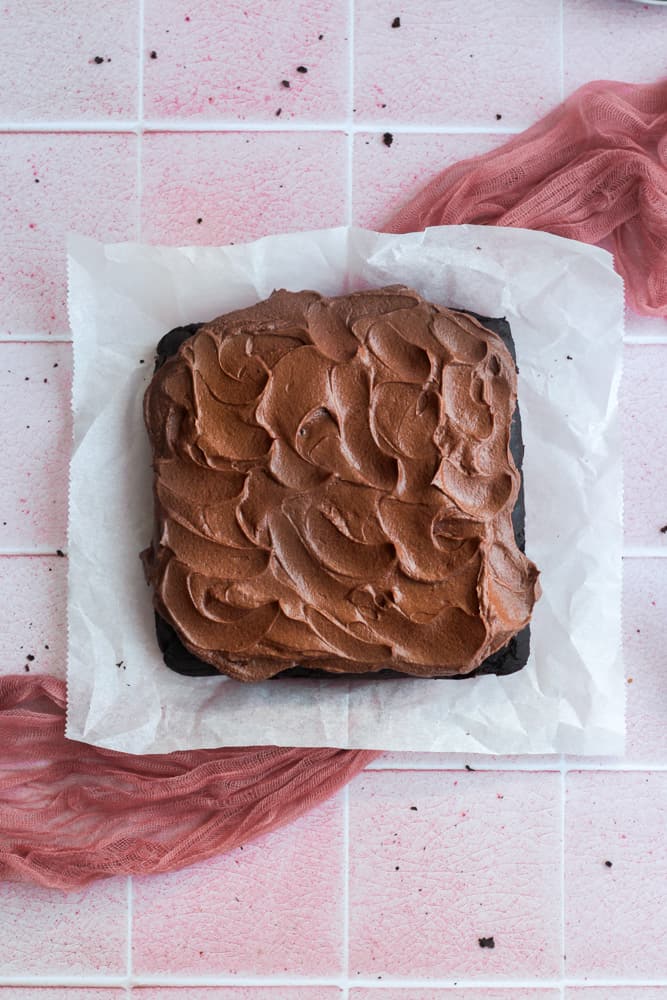 gluten free chocolate snack cake topped with chocolate buttercream on parchment paper on a pink backdrop