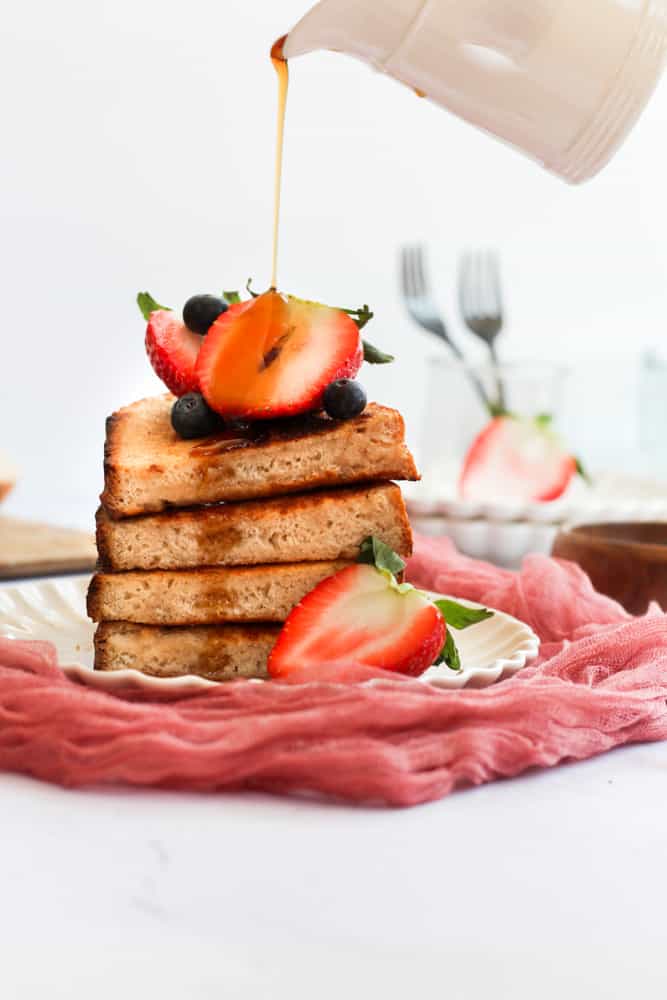 4 slices of sourdough French toast stacked on a white plate and topped with cut berries. maple syrup is being poured over the stack