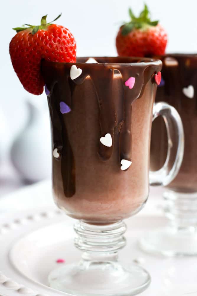 strawberry hot chocolate in a clear mug with a dripped chocolate rim and heart sprinkles. The glass is finished with a cut strawberry