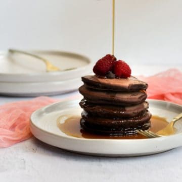 chocolate pancakes topped with raspberries on a white plate with maple syrup being poured over them