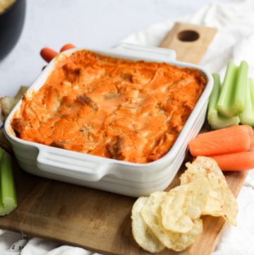 dairy free buffalo chicken dip baked in a white baking dish on a wood swuare board lined with carrots, celery, and potato chips on a whit backdrop