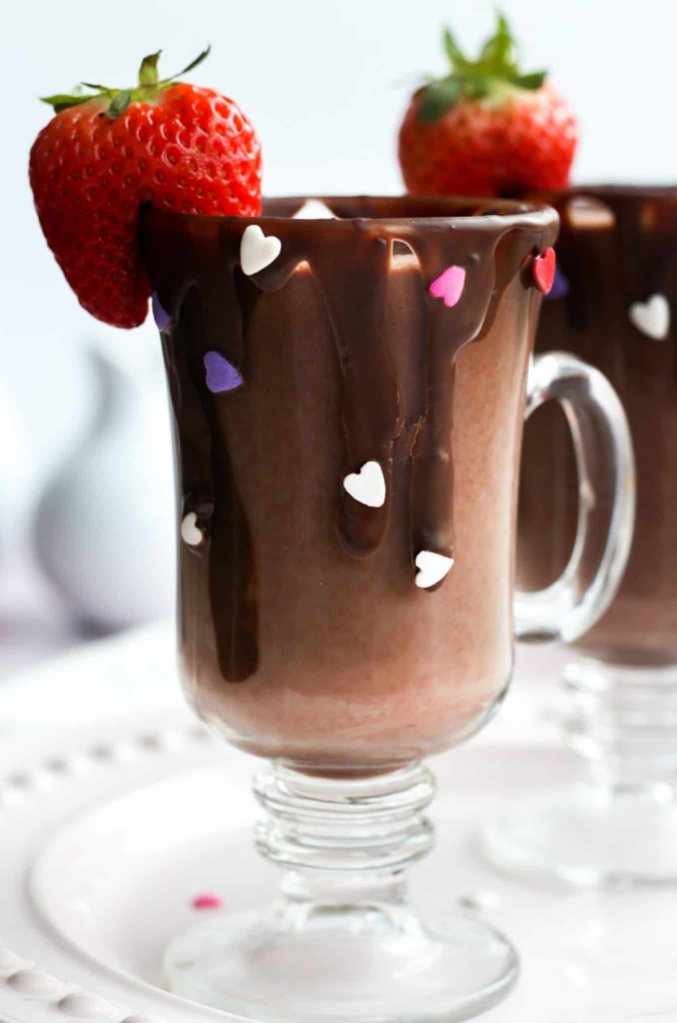 strawberry hot chocolate in a clear mug with a dripped chocolate rim and heart sprinkles. The glass is finished with a cut strawberry