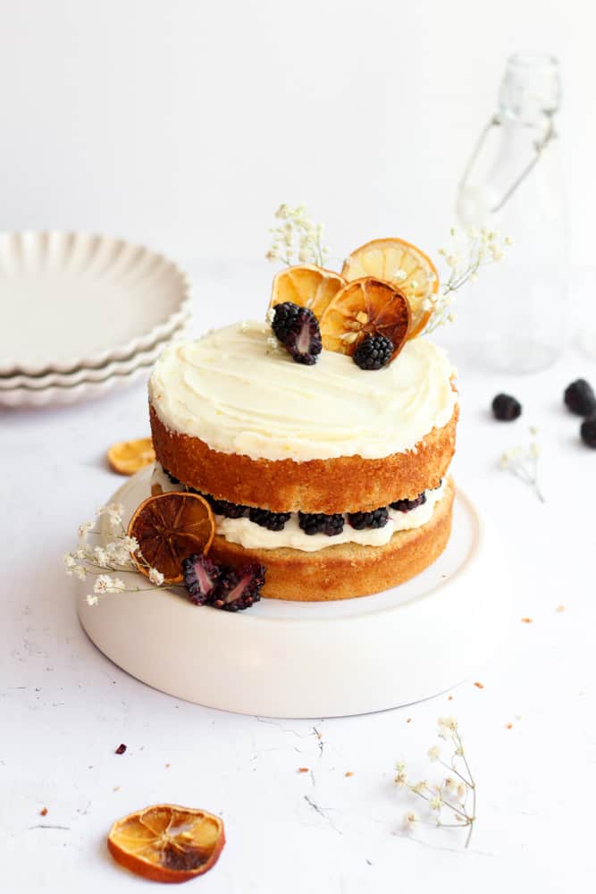 naked lemon and blackberry cake on a white cake stand decorated with dried lemons and flowers