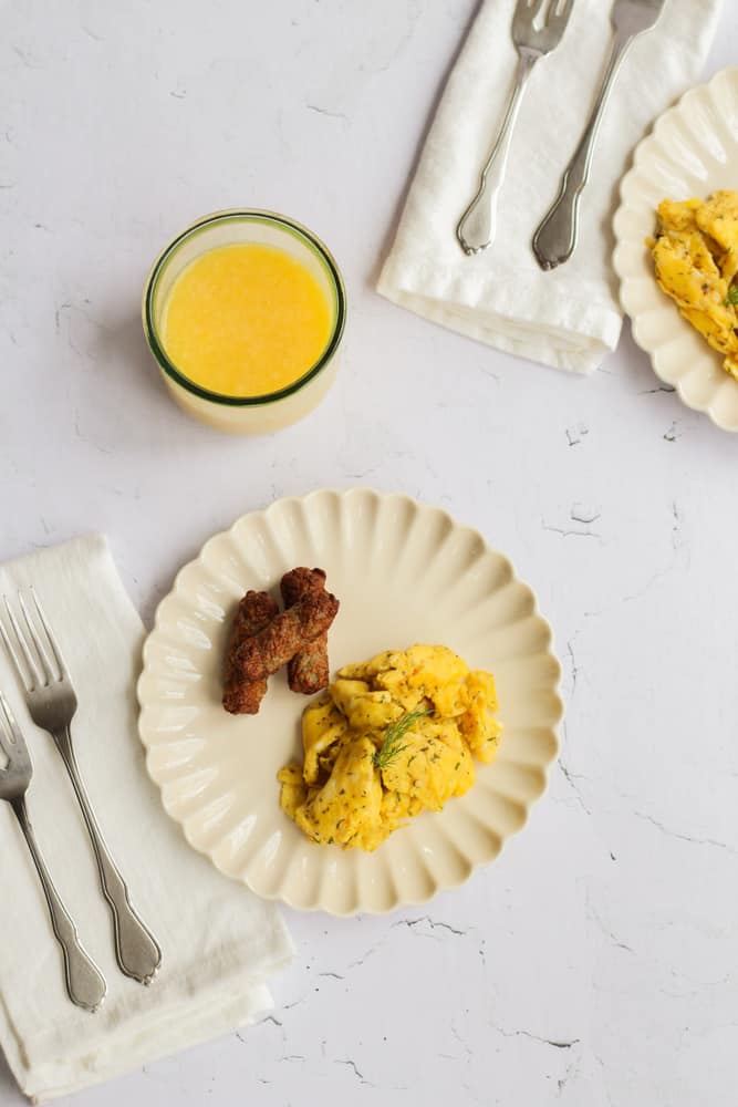 Scrambled eggs with sausage on a white plate with orange juice and silverware around it.