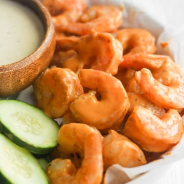 buffalo shrimp in a white bowl with cucumbers and dip.