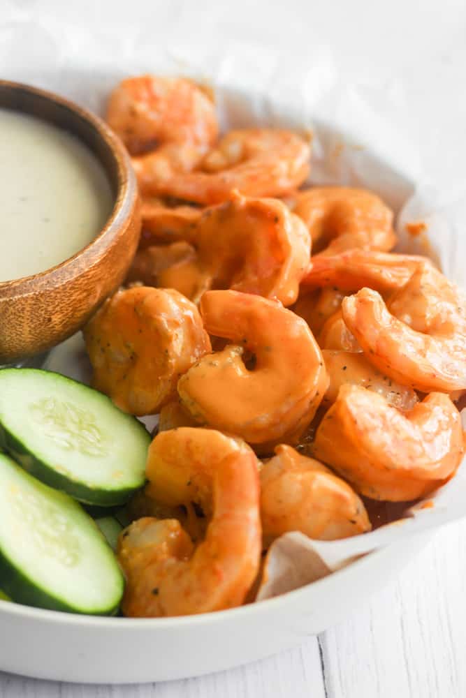 buffalo shrimp in a white bowl with cucumbers and dip.