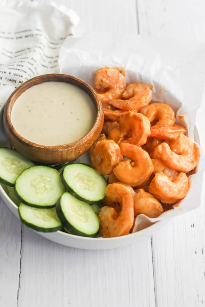 healthy buffalo shrimp in a white bowl with cucumber slices and dip.