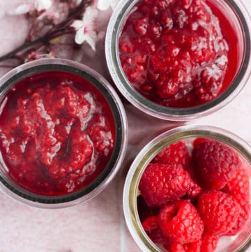three glass jars on a pink backdrop. two have raspberry compote, one has fresh raspberries.