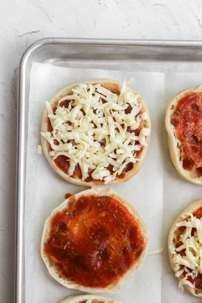 english muffin pizzas topped with shredded cheese on parchment paper.
