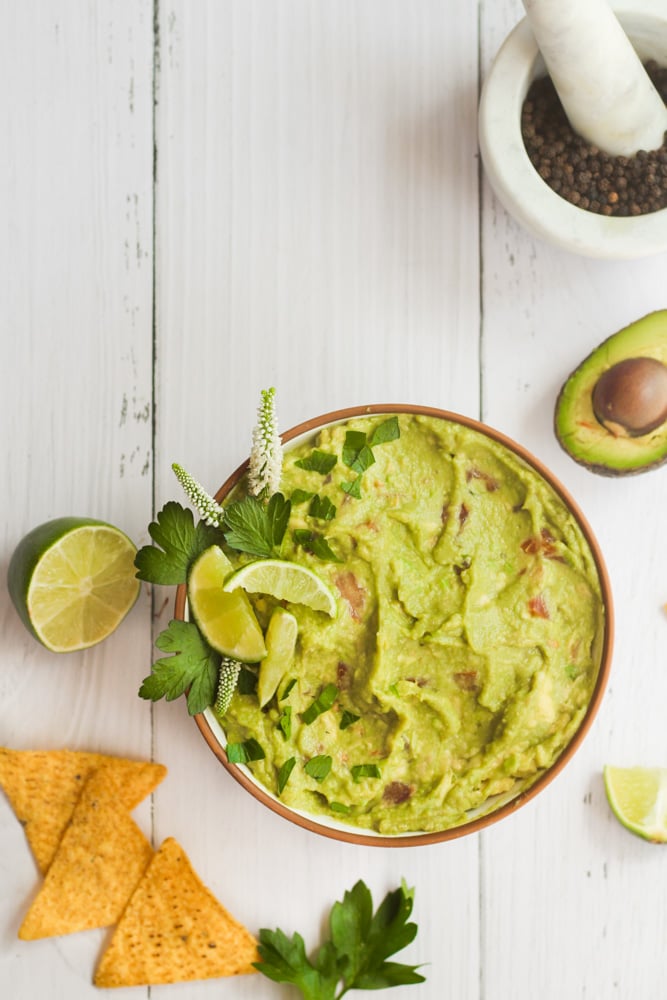 easy guacamole in a white bowl, tortilla chips, limes, and an avocado are around it.