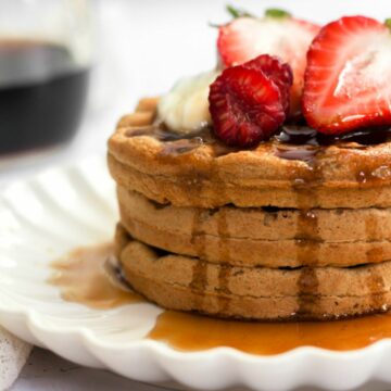 3 oat flour waffles stacked on a white plate topped with strawberries and maple syrup.