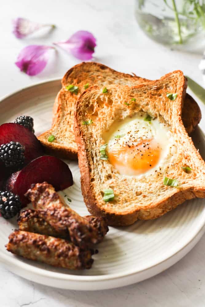 air fryer egg in a hole on a white plate with sausage, fruit, and herbs.