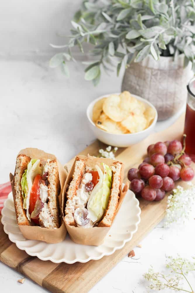 Panera's chicken salad recipe on a sandwich wrapped in paper on plate with grapes and chips.