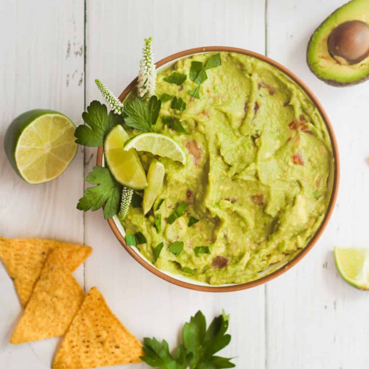 guacamole recipe with no onion in a white bowl with chips, limes, and a half an avocado surrounding it.