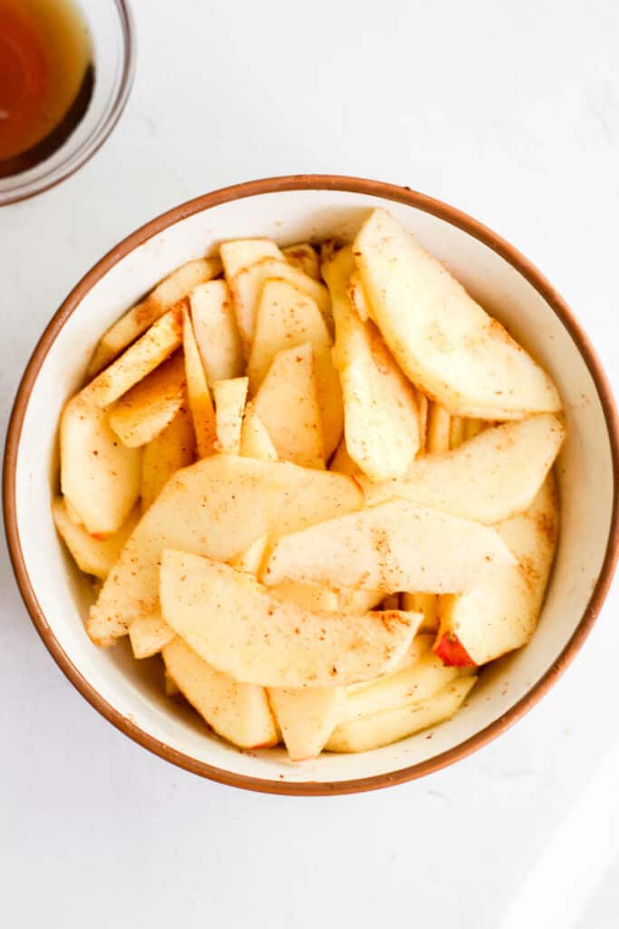 slices of apples in white bowl with cinnamon, butter, and maple syrup.