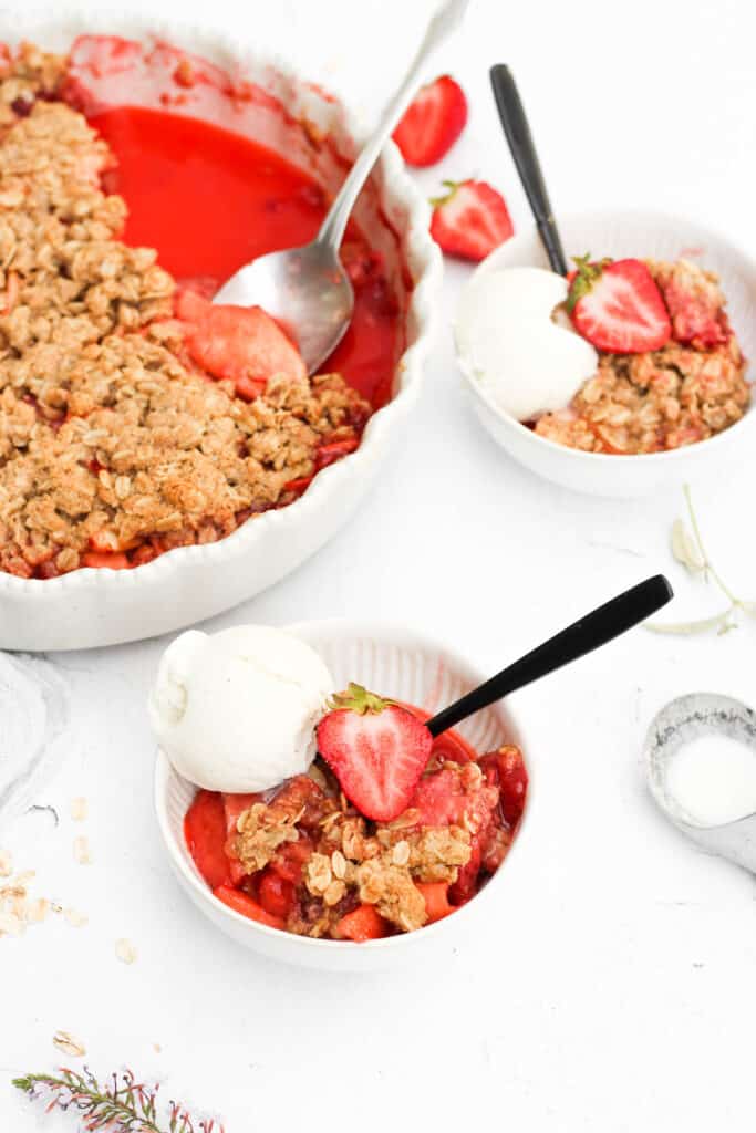 healthy strawberry crisp in two white bowls with scoops of vanilla ice cream and strawberries.