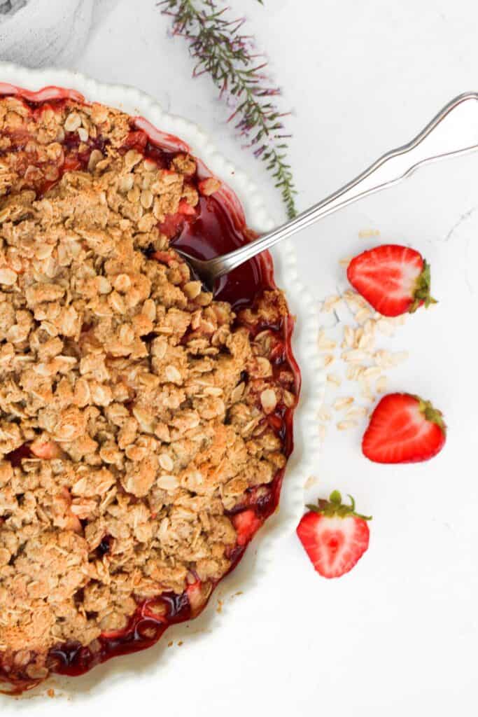 apple strawberry crumble in pie plate with metal spoon and halved strawberries.