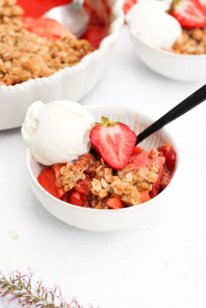apple strawberry crisp in a white bowl with a scoop of vanilla ice cream and halved strawberry.