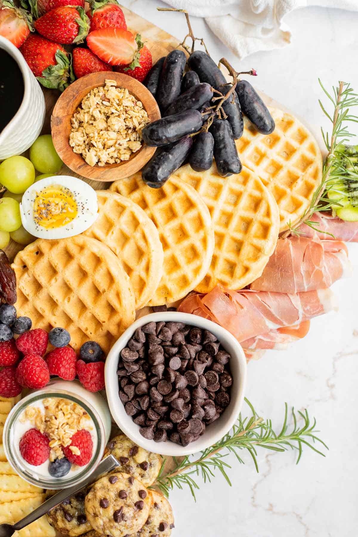 mini waffles, meat, chocolate chips, fruit, and yogurt on a brunch board.
