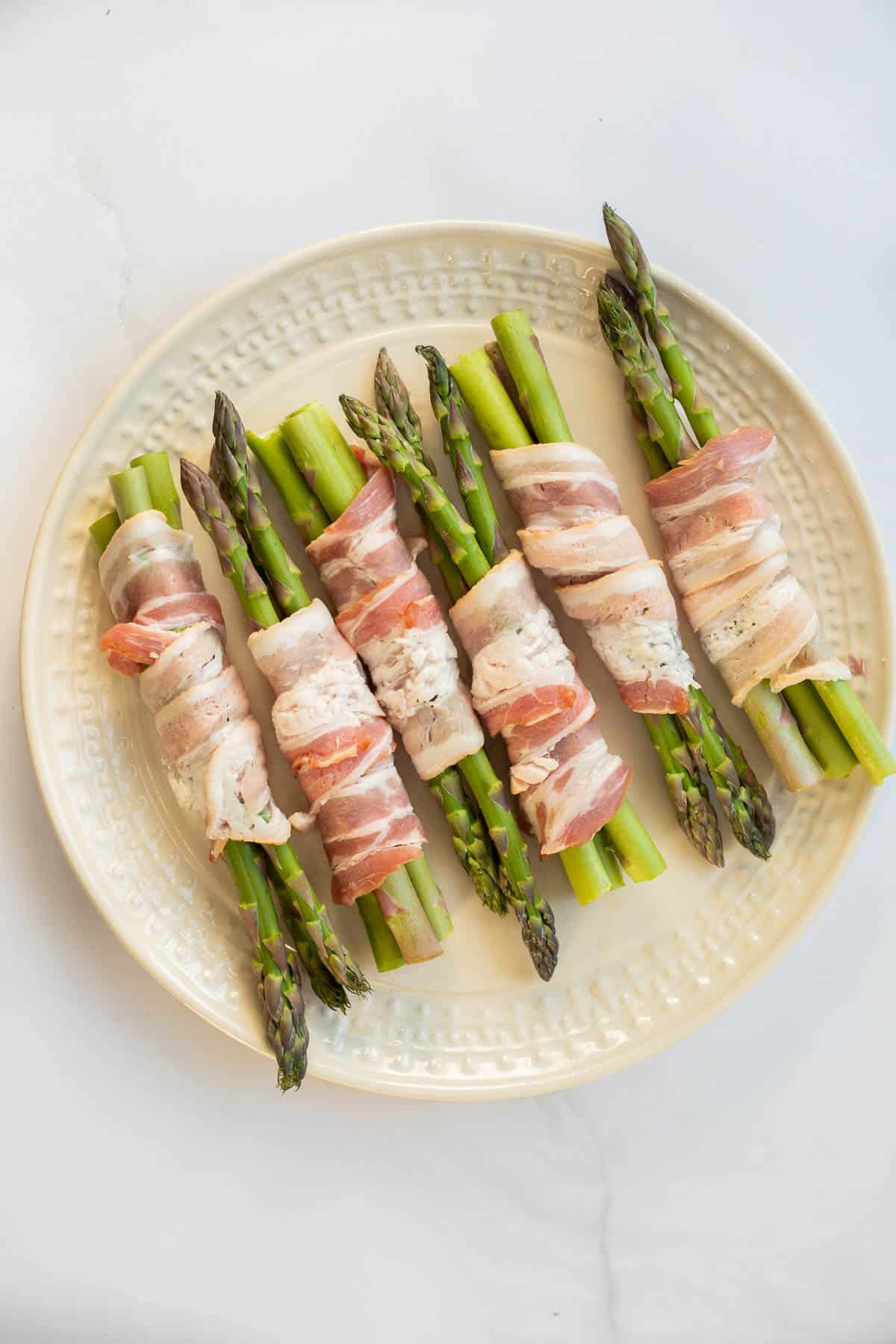 asparagus spears wrapped in raw bacon in a white plate.
