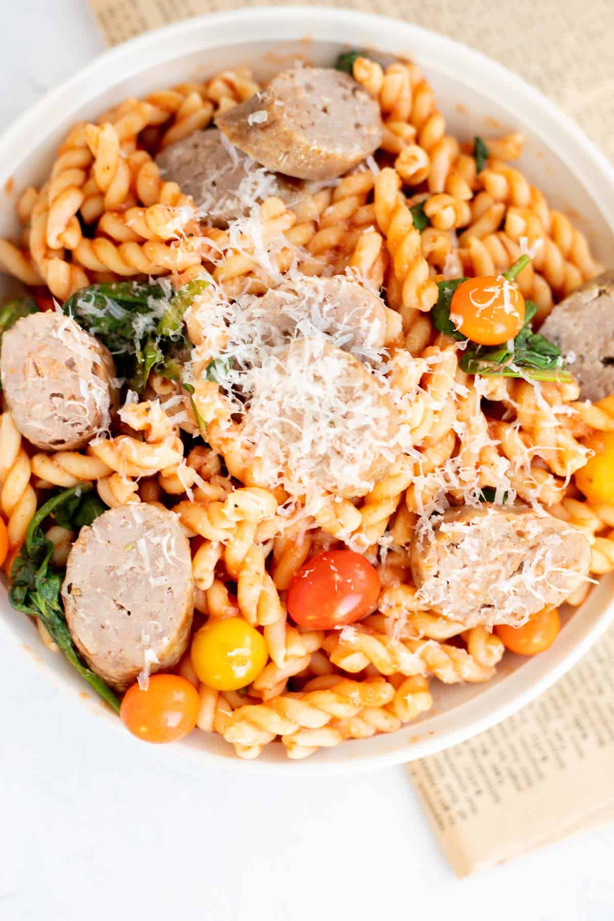 white bowl of pasta with spinach, tomatoes, cheese, and slices of sausage.