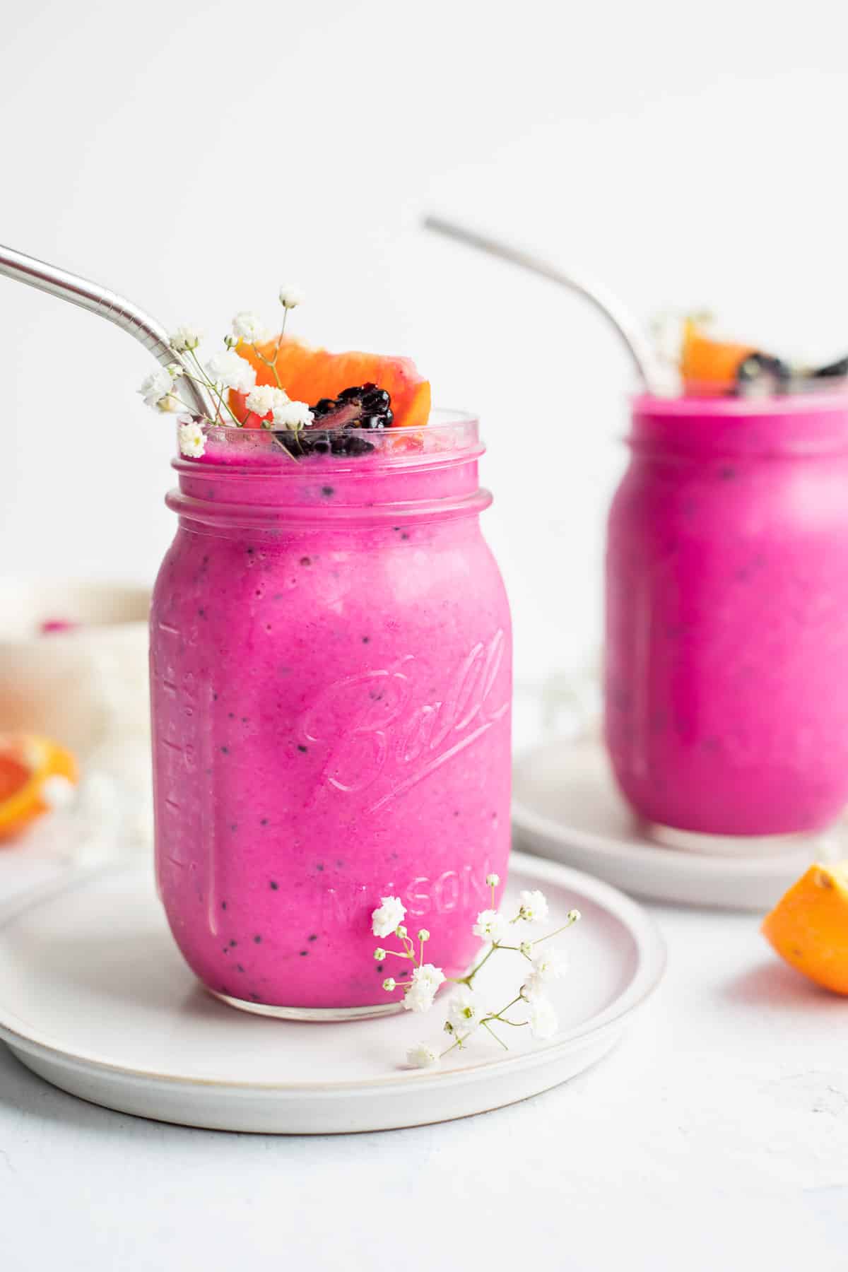 two dragon fruit smoothies in glass jars with straws topped with blackberries and flowers.